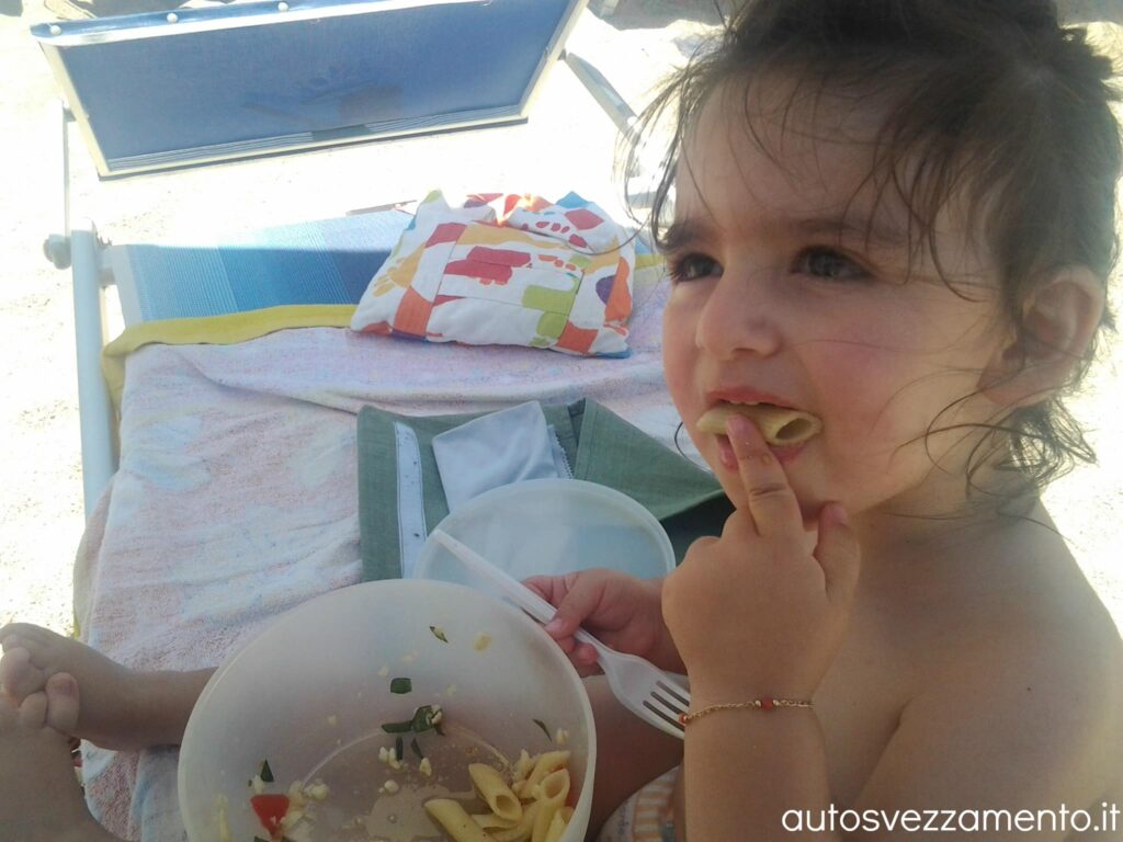 Bambina si svezza da sola in spiaggia mangiando con le mani un bel piatto di pasta